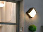 Applique murale exterieur LED imperméable et moderne installé sur un mur avec une porte vitrée à gauche et une plante verte en dessous