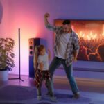 Un homme et une petite fille dans un salon qui dansent, derrière eux se trouve une TV au mur, une baffle et le lampadaire LED d'ambiance intelligent allumé.