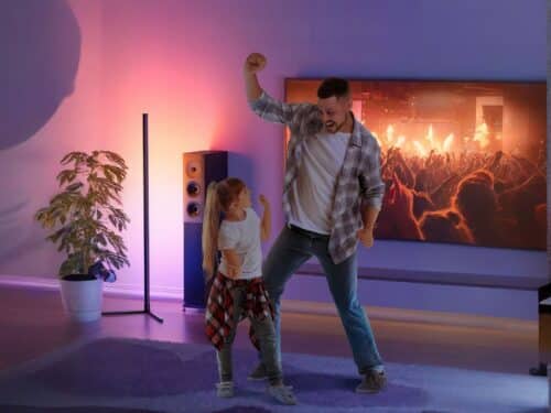Un homme et une petite fille dans un salon qui dansent, derrière eux se trouve une TV au mur, une baffle et le lampadaire LED d'ambiance intelligent allumé.