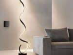 Lampadaire LED noir, design moderne en spirale, allumé et posé à côté d'un canapé gris foncé.