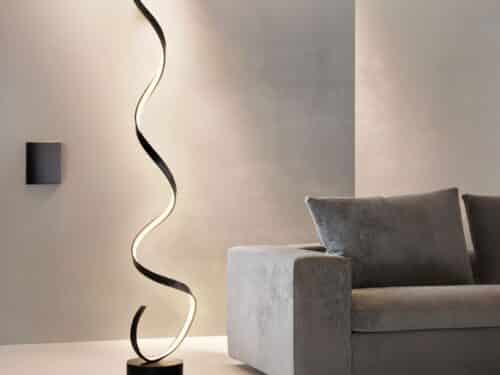 Lampadaire LED noir, design moderne en spirale, allumé et posé à côté d'un canapé gris foncé.