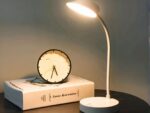 Lampe à poser LED blanche design sur pied, à côté d'un livre et une horloge posés sur une table.