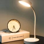 Lampe à poser LED blanche design sur pied, à côté d'un livre et une horloge posés sur une table.