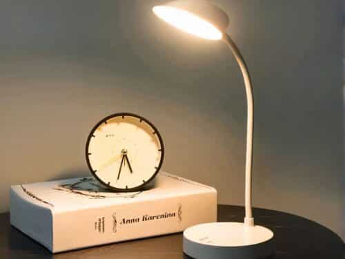 Lampe à poser LED blanche design sur pied, à côté d'un livre et une horloge posés sur une table.