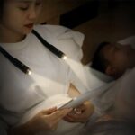 Liseuse LED de cou flexible et polyvalente Femme dans son lit portant la liseuse Led de cou, allumée et lisant un livre dans le noir.