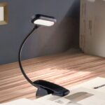 Liseuse LED noire flexible à pince Liseuse LED noire à pince avec tige flexible, allumée et accrochée à un livre. Le tout sur un bureau en bois.
