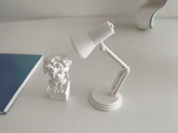 Liseuse LED mini, toute blanche, sur socle, posé sur un bureau blanc et face à une mini tête statue blanche.