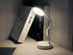 Liseuse LED sur socle rond, avec tige flexible, allumée et posé sur un bureau à côté de livres.