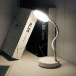 Liseuse LED rechargeable avec tige flexible Liseuse LED sur socle rond, avec tige flexible, allumée et posé sur un bureau à côté de livres.