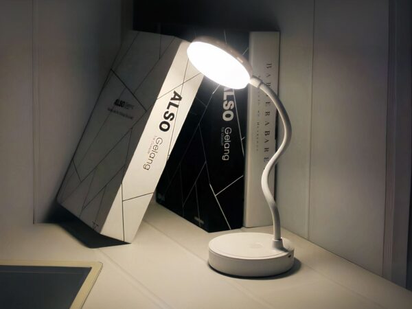 Liseuse LED sur socle rond, avec tige flexible, allumée et posé sur un bureau à côté de livres.