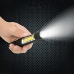 Mini lampe led multifonctionnelle étanche et noire Mini lampe led multifonctionnelle étanche et noire, tenue dans une main, projetant une lumière blanche, sur un fond noir.