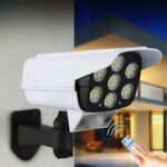 Projecteur LED solaire et télécommandé en forme de caméra Projecteur LED solaire et télécommandé en forme de caméra installé sur un mur avec une maison en fond et la main d'une personne qui tient une télécommande en bas à droite