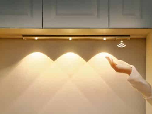 Réglette LED de cuisine, allumée et installée sous un meuble haut de cuisine. Une main passe pour déclencher le détecteur de mouvement.