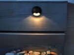 Spot LED extérieur noir et arrondi allumé et installé contre un mur en bois.