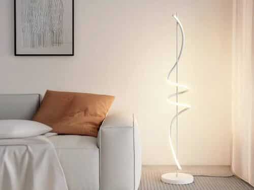 Lampadaire LED fer créatif spirale blanc allumé et installé à côté d'un canapé beige avec un coussin marron.