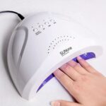 Lampe LED ongle pour vernis simple ou UV, allumée et utilisée par une mannequin, on ne voit que la machine et la main.