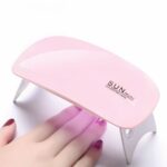 Lampe LED ongle UV pliable et portable Lampe LED ongles UV pliable et portable, rose et blanche, allumée avec la main d'une femme qui sèche son vernis.