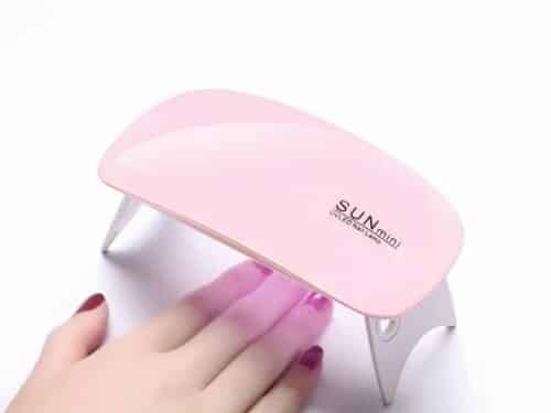 Lampe LED ongles UV pliable et portable, rose et blanche, allumée avec la main d'une femme qui sèche son vernis.