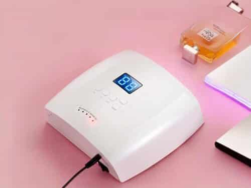 Lampe LED ongle UV rechargeable, de dos sur une table rose avec un parfum à côté.