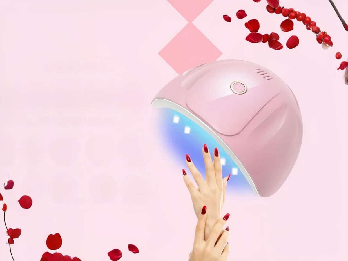 Lampe LED ongle UV rose au design arrondi, allumée et mise en scène sur un fond rose avec des pétales de roses rouges et deux mains d'une femme avec les ongles vernis en rouge.