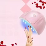 Lampe LED ongle UV rose design arrondi Lampe LED ongle UV rose au design arrondi, allumée et mise en scène sur un fond rose avec des pétales de roses rouges et deux mains d'une femme avec les ongles vernis en rouge.