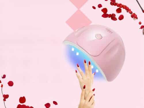 Lampe LED ongle UV rose au design arrondi, allumée et mise en scène sur un fond rose avec des pétales de roses rouges et deux mains d'une femme avec les ongles vernis en rouge.