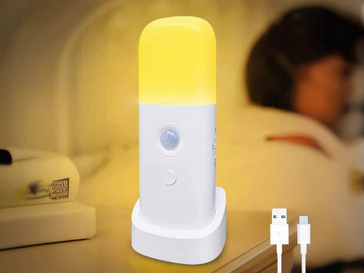 Lampe LED rechargeable à détecteur de mouvement posée sur une table de nuit avec une personne allongée dans un lit en fond