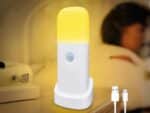 Lampe LED rechargeable à détecteur de mouvement posée sur une table de nuit avec une personne allongée dans un lit en fond