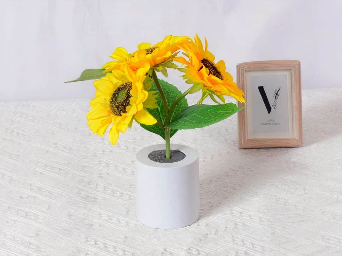 Lampe LED rechargeable effet pot de fleurs posée sur une table avec une nappe blanche et un cadre en fond