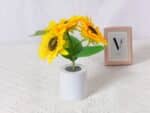 Lampe LED rechargeable effet pot de fleurs posée sur une table avec une nappe blanche et un cadre en fond