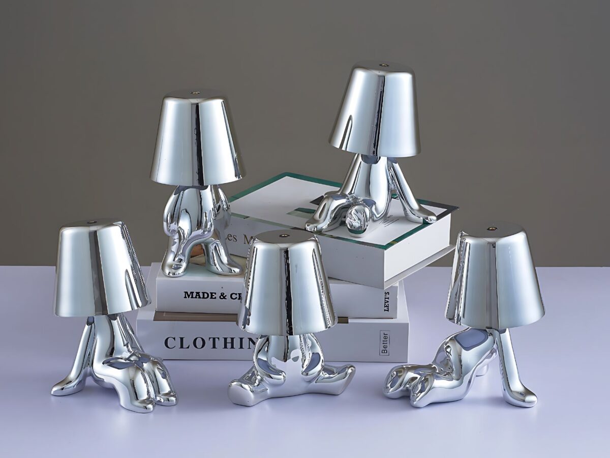 5 lampes veilleuses tactiles argentées en exposition sur une étagère et une pile de livres. Chacune représente un petit bonhomme dans une posture originale. Trois sont assis de façon différente, un est debout et le dernier est à quatre pattes.