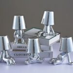 Lampes LED veilleuses tactiles argentées 5 lampes veilleuses tactiles argentées en exposition sur une étagère et une pile de livres. Chacune représente un petit bonhomme dans une posture originale. Trois sont assis de façon différente, un est debout et le dernier est à quatre pattes.