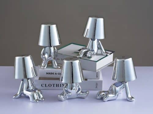 5 lampes veilleuses tactiles argentées en exposition sur une étagère et une pile de livres. Chacune représente un petit bonhomme dans une posture originale. Trois sont assis de façon différente, un est debout et le dernier est à quatre pattes.