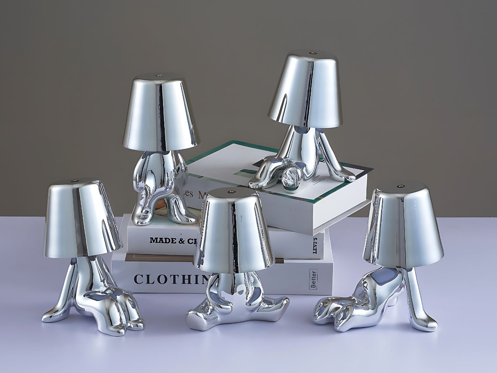 5 lampes veilleuses tactiles argentées en exposition sur une étagère et une pile de livres. Chacune représente un petit bonhomme dans une posture originale. Trois sont assis de façon différente, un est debout et le dernier est à quatre pattes.