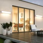 Applique murale LED extérieure imperméable et solaire 2 appliques murales LED imperméables entourent une baie vitrée donnant dans une salle à manger. Elles éclairent une terrasse en bois, décorée de 3 pots de fleurs et d'un salon de jardin blanc.