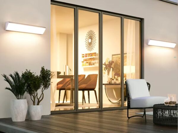 2 appliques murales LED imperméables entourent une baie vitrée donnant dans une salle à manger. Elles éclairent une terrasse en bois, décorée de 3 pots de fleurs et d'un salon de jardin blanc.