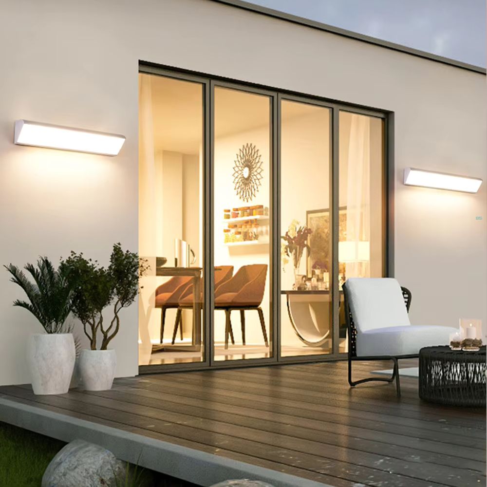 Applique murale LED extérieure imperméable et solaire 2 appliques murales LED imperméables entourent une baie vitrée donnant dans une salle à manger. Elles éclairent une terrasse en bois, décorée de 3 pots de fleurs et d'un salon de jardin blanc.