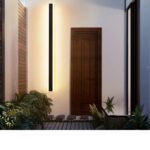 Applique murale verticale LED étanche Un pas de porte éclairé par une applique murale verticale LED étanche qui diffuse un lumière blanche chaude. L'applique est positionnée à gauche de la porte. Devant elle deux buissons.