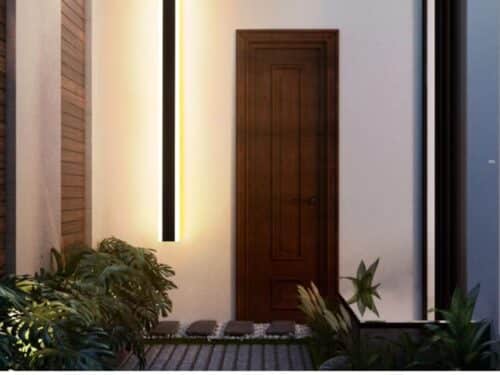 Un pas de porte éclairé par une applique murale verticale LED étanche qui diffuse un lumière blanche chaude. L'applique est positionnée à gauche de la porte. Devant elle deux buissons.