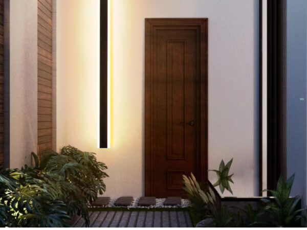 Un pas de porte éclairé par une applique murale verticale LED étanche qui diffuse un lumière blanche chaude. L'applique est positionnée à gauche de la porte. Devant elle deux buissons.