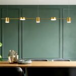 Le lustre à led doré au design moderne au-dessus d'une table de salle à manger en bois au fond un mur vert