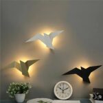 Applique murale LED en forme d'oiseau Les trois modèles d'appliques murales LED en forme d'oiseau doré, blanc et noir au-dessus d'un bureau avec une horloge, sur un mur taupe.