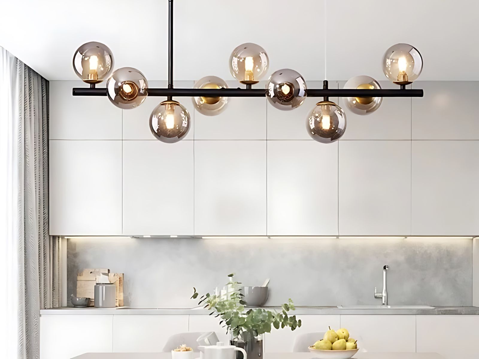 Le lustre suspension en verre au style contemporain dans une cuisine au dessus d'une grande table blanche dessus avec une plante verte et un saladier avec des citrons jaunes