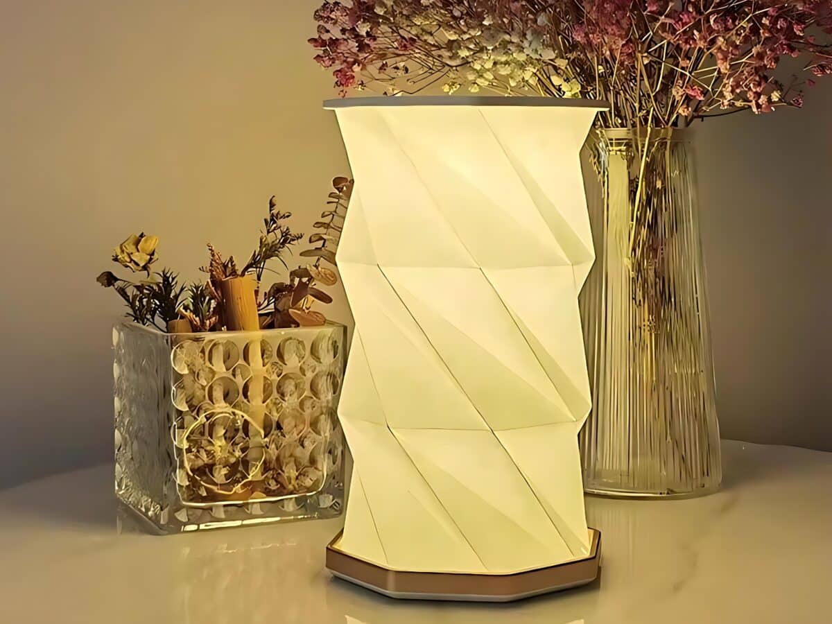 IMG_lampe_de_chevet_LED_pliable_au_design_d_origami_1 Lampe de chevet LED pliable au design d'origami blanc. La lampe est allumée et posée devant un vase transparent avec des fleurs violettes et un récipient rectangulaire transparent avec des herbes et fleurs sèches.