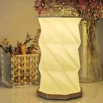 Lampe de chevet LED pliable au design d'origami Lampe de chevet LED pliable au design d'origami blanc. La lampe est allumée et posée devant un vase transparent avec des fleurs violettes et un récipient rectangulaire transparent avec des herbes et fleurs sèches.