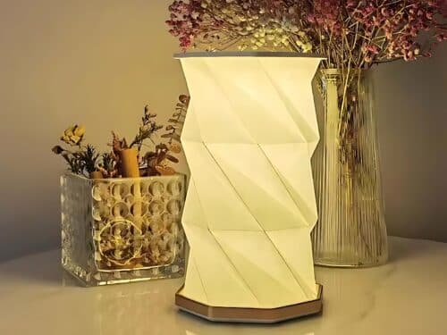 Lampe de chevet LED pliable au design d'origami blanc. La lampe est allumée et posée devant un vase transparent avec des fleurs violettes et un récipient rectangulaire transparent avec des herbes et fleurs sèches.