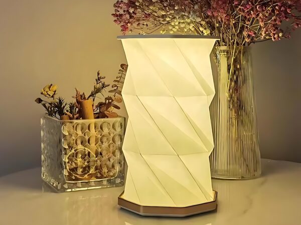 Lampe de chevet LED pliable au design d'origami blanc. La lampe est allumée et posée devant un vase transparent avec des fleurs violettes et un récipient rectangulaire transparent avec des herbes et fleurs sèches.