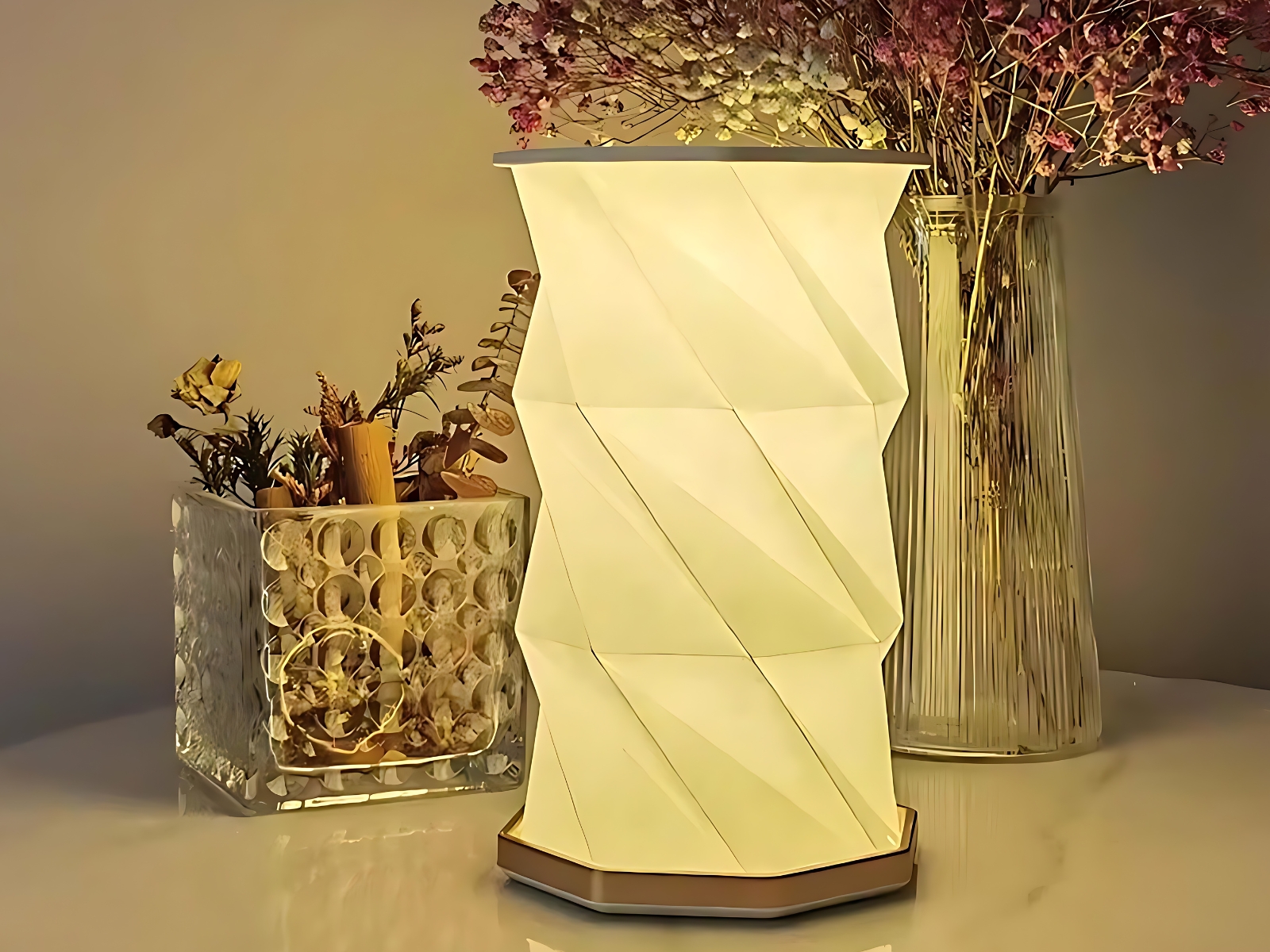 Lampe de chevet LED pliable au design d'origami Lampe de chevet LED pliable au design d'origami blanc. La lampe est allumée et posée devant un vase transparent avec des fleurs violettes et un récipient rectangulaire transparent avec des herbes et fleurs sèches.