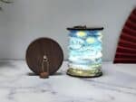 Lampe de chevet LED pliable style lanterne japonaise aux motifs bleus et verts. Pliable en forme accordéon. Les deux extrémités sont en bois foncé. Une lampe pliée se trouve à gauche de la lampe ouverte et allumée. Elles sont posées sur une surface type marbre blanc. Un mur gris en fond.