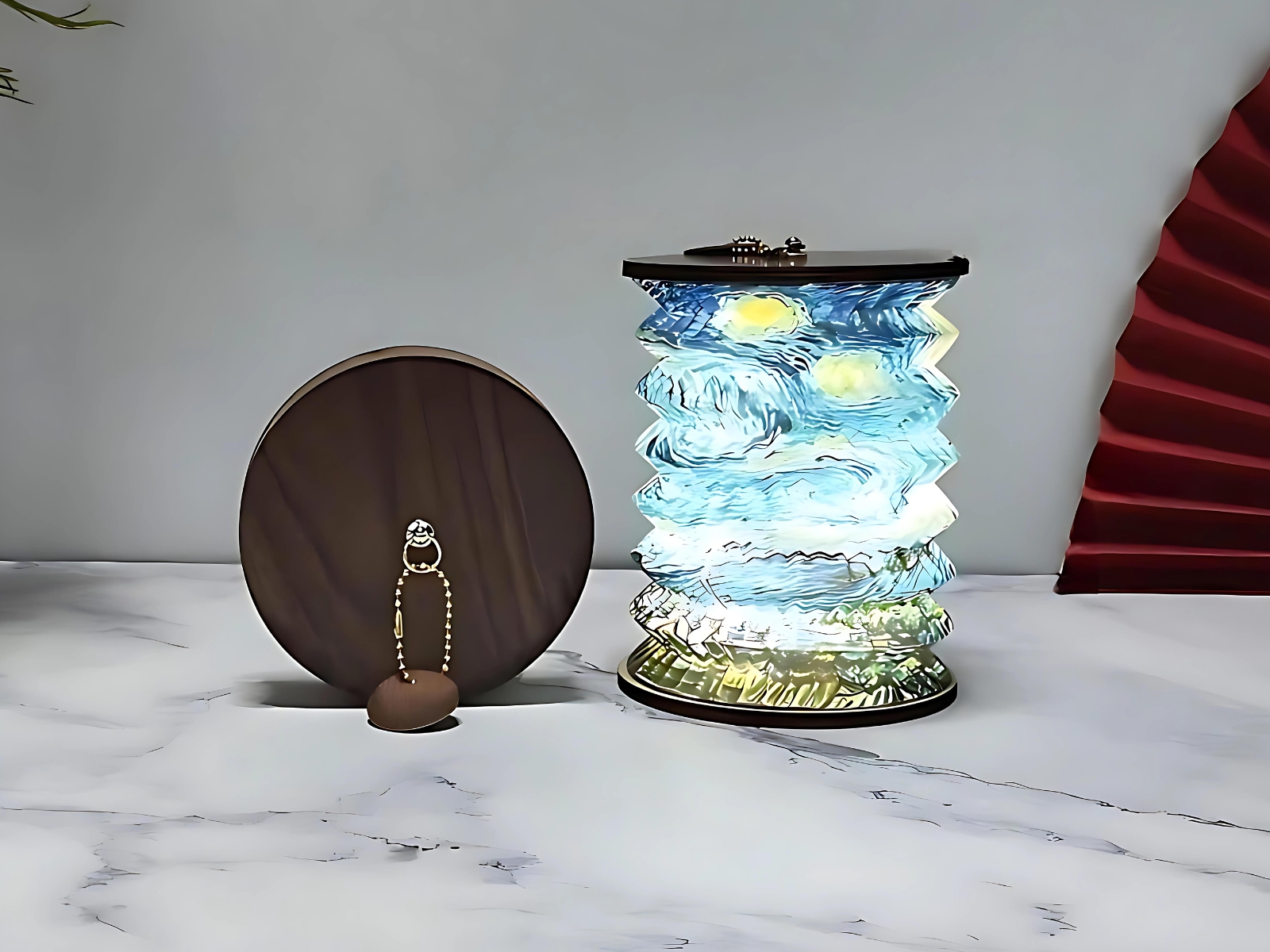 Lampe de chevet LED pliable style lanterne japonaise aux motifs bleus et verts. Pliable en forme accordéon. Les deux extrémités sont en bois foncé. Une lampe pliée se trouve à gauche de la lampe ouverte et allumée. Elles sont posées sur une surface type marbre blanc. Un mur gris en fond.